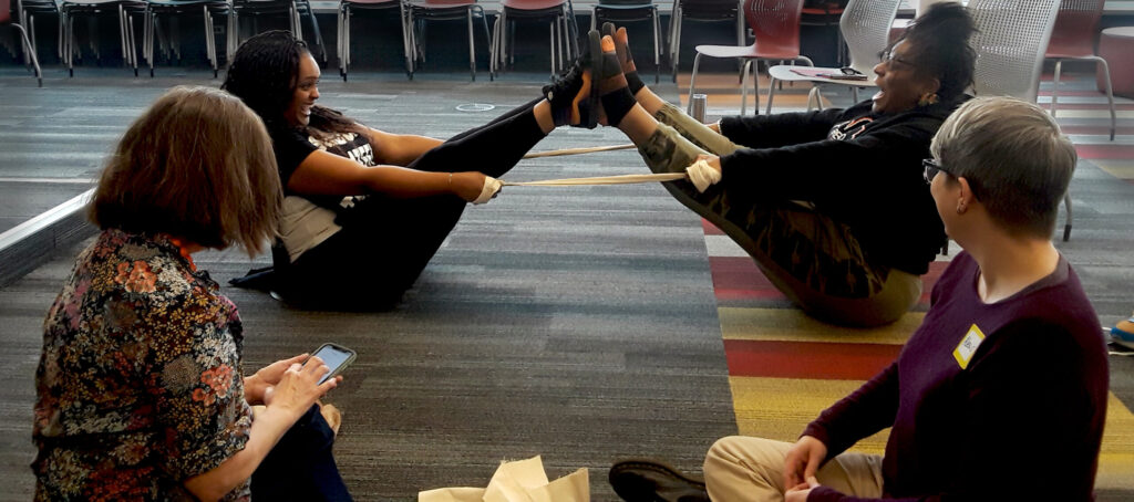 Two participants are seated facing each other, leaning back and balancing feet-to-feet with a resistance band or fabric strap looped between their raised legs and hands — a partner balance/trust exercise