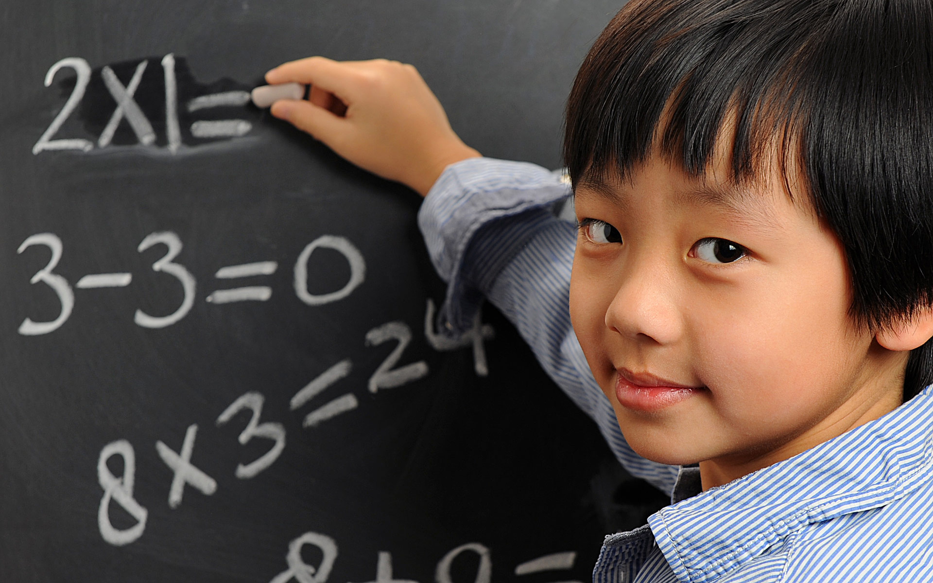 A boy at the blackboard writing math equations and looking back at the camera